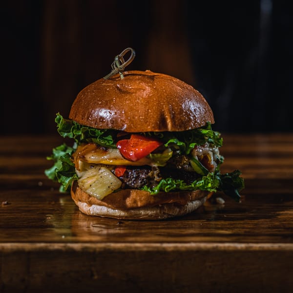 A BB's Burger Factory burger with lettuce, cheese, red peppers, and onions on a wooden surface.